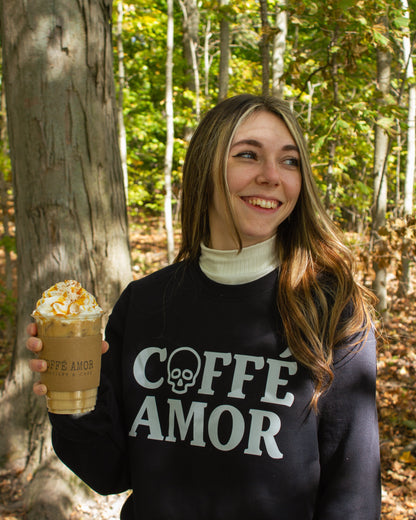 A woman standing in a wooded outdoor setting, holding a coffee drink and wearing a black sweatshirt with the words 'COFFE AMOR' with a skull graphic as the O in "COFFE".