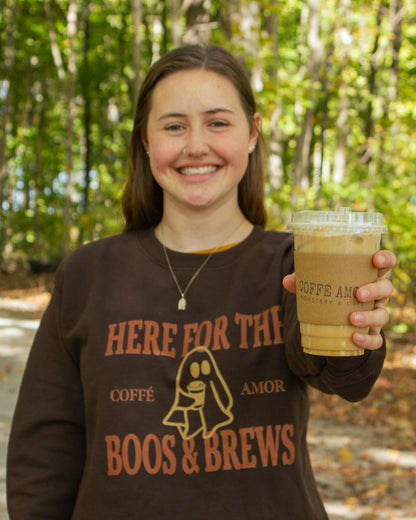 Woman holding a coffee and wearing a brown crewneck that says "Here for the boos & brews" with a ghost graphic holding a coffee in the middle.