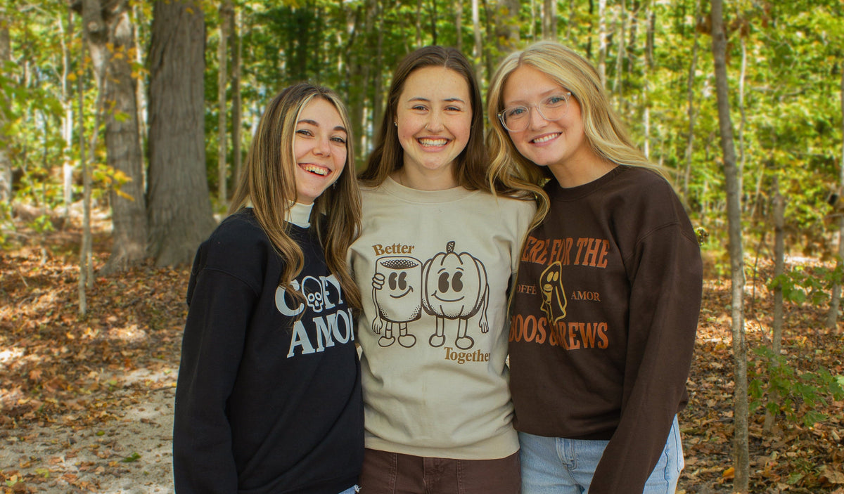 Three young women standing together in a wooded area, each wearing fall coffe amor crewnecks.